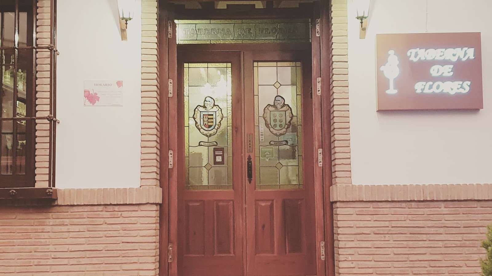 Fachada encalada de La Taberna de Flores en Colmenar con el nombre del restaurante en la pared junto a la puerta de cristal